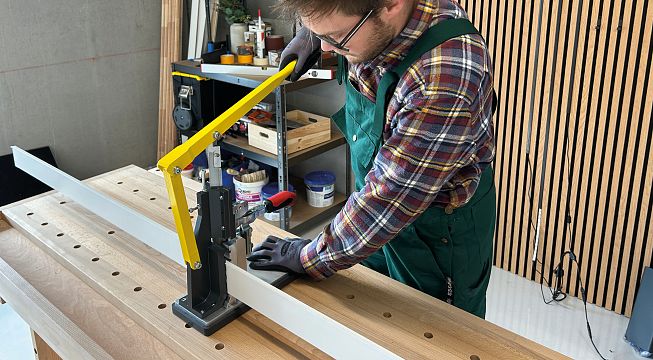 Ein Handwerker in kariertem Hemd und grüner Latzhose bedient eine manuelle Schneidemaschine auf einer Werkbank. Verschiedene Werkzeuge sind im Hintergrund.