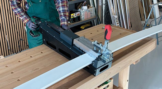 Person arbeitet in einer Werkstatt und benutzt eine Holzbearbeitungsmaschine mit einem roten Griff an einem stabilen Holztisch.