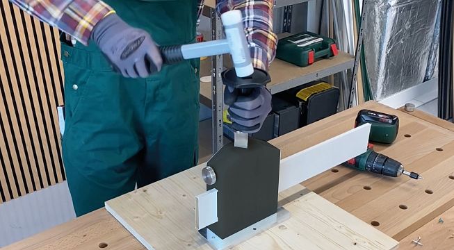 Ein Handwerker trägt Handschuhe und befestigt ein Metallteil mit einem Schraubenzieher in einer Werkstatt. Im Hintergrund sind Werkzeuge und Regale zu sehen.