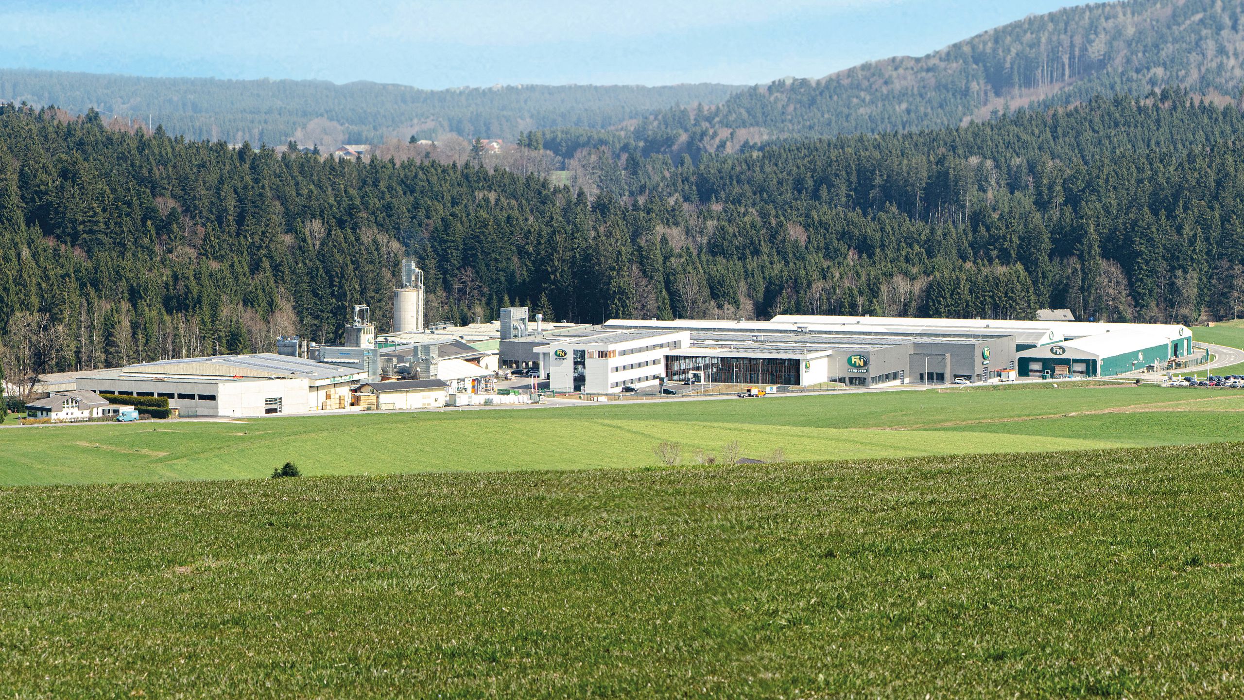 Rolling green fields leading up to a large industrial facility surrounded by dense forest and hills under a clear blue sky.